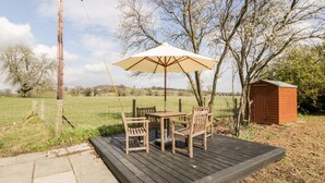 A wooden deck with a table and chairs, an umbrella, and a small shed in a rural setting.