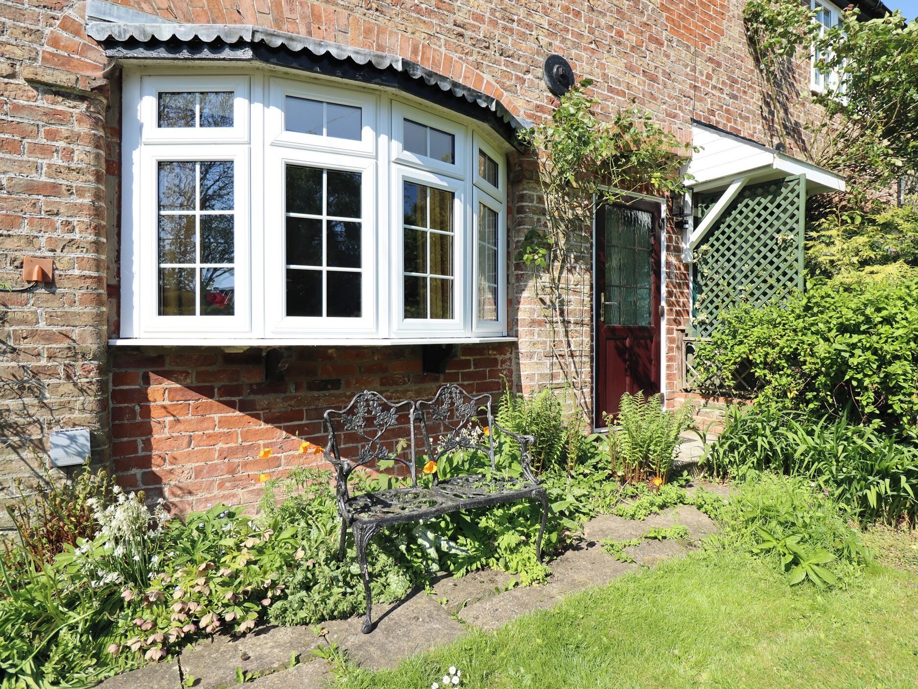 A brick house with a white window, a red door, and a garden with a bench.