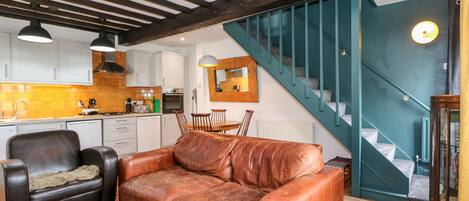 A modern kitchen with white cabinets, a dining area with a brown leather sofa, and a staircase with teal railings.