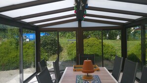 Un patio cubierto con una mesa y sillas, una lámpara de pie de terracota y vistas a un jardín con arbustos y una estructura azul al fondo.