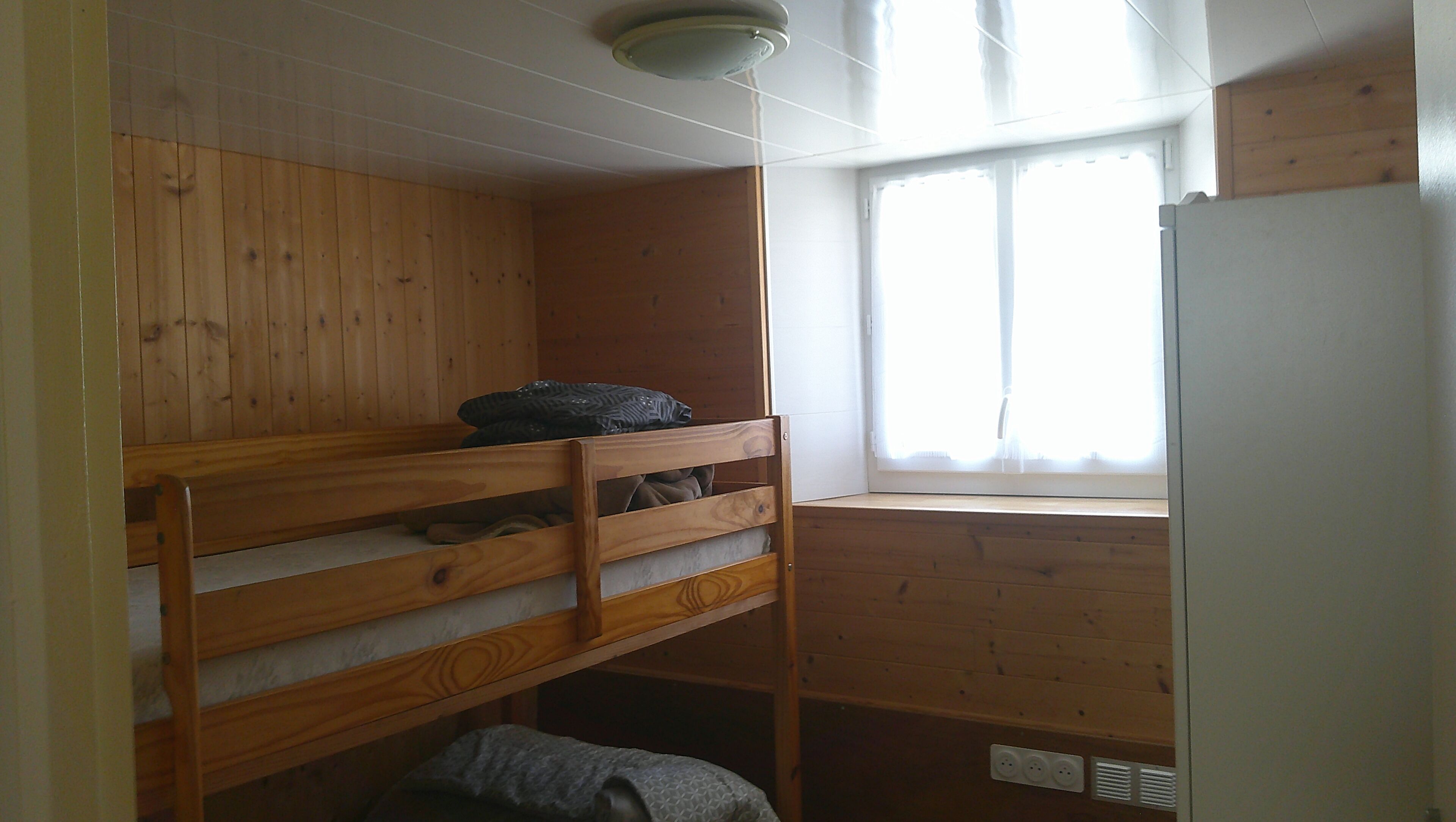 Ein kleiner, kompakter Raum mit Etagenbett, einem Fenster mit Jalousien und einem integrierten Kühlschrank.