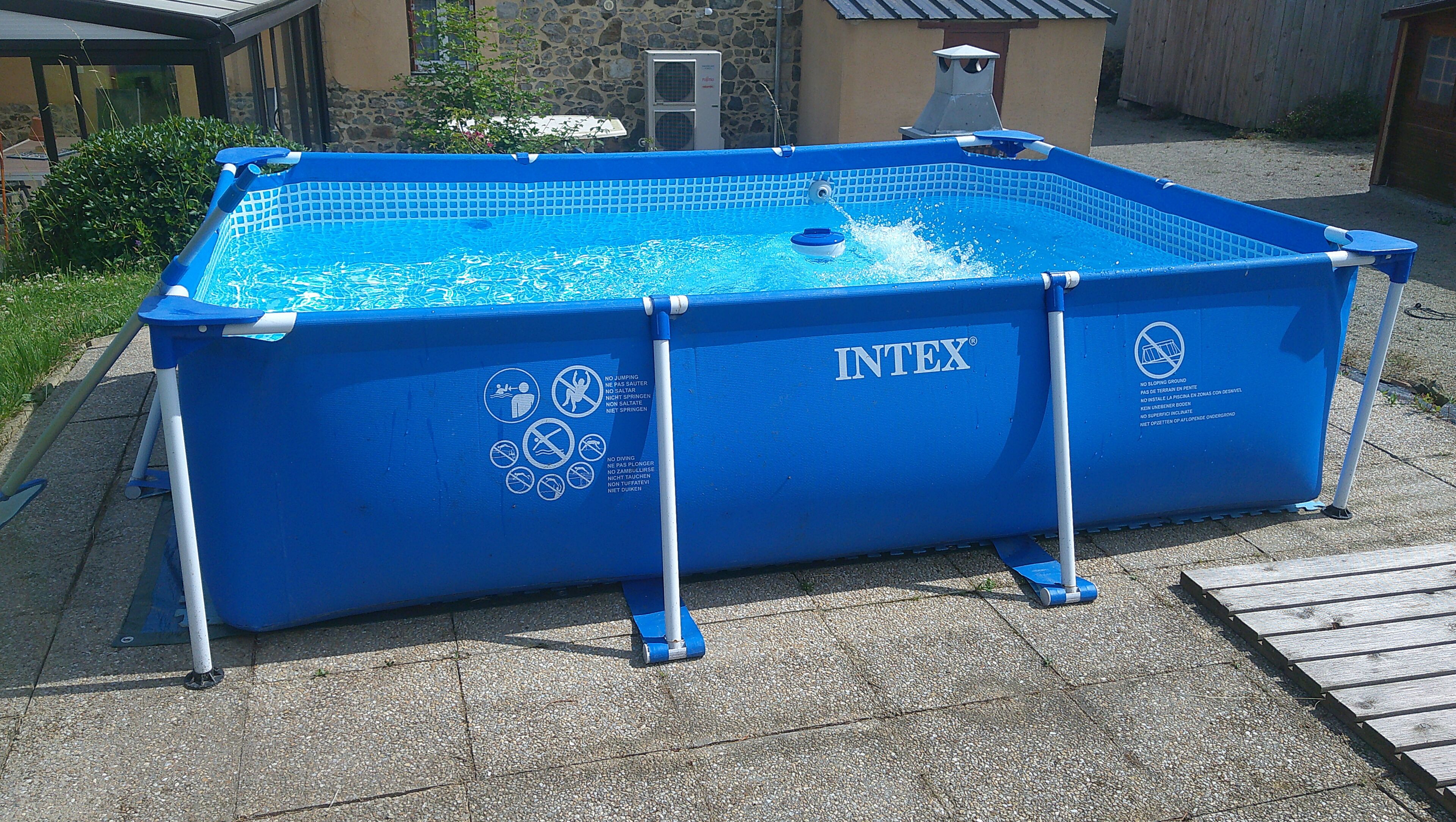 A blue above-ground pool with INTEX branding, surrounded by a concrete deck and a stone wall in the background.