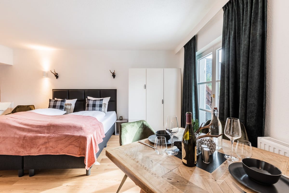 A modern hotel room with a bed, a wooden table with wine glasses and bottles, a green chair, and a white wardrobe.