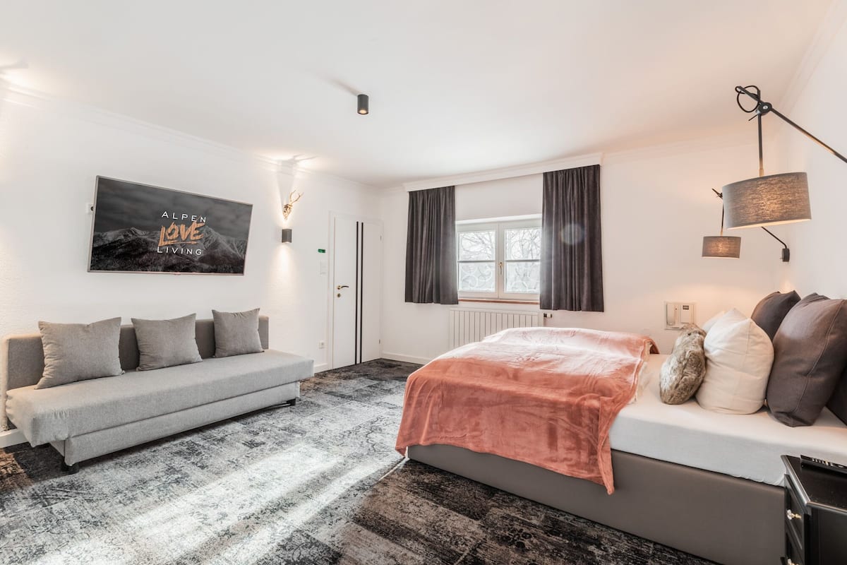 A modern bedroom with a grey sofa, a bed with a pink blanket, a wall-mounted lamp, and a window with curtains.