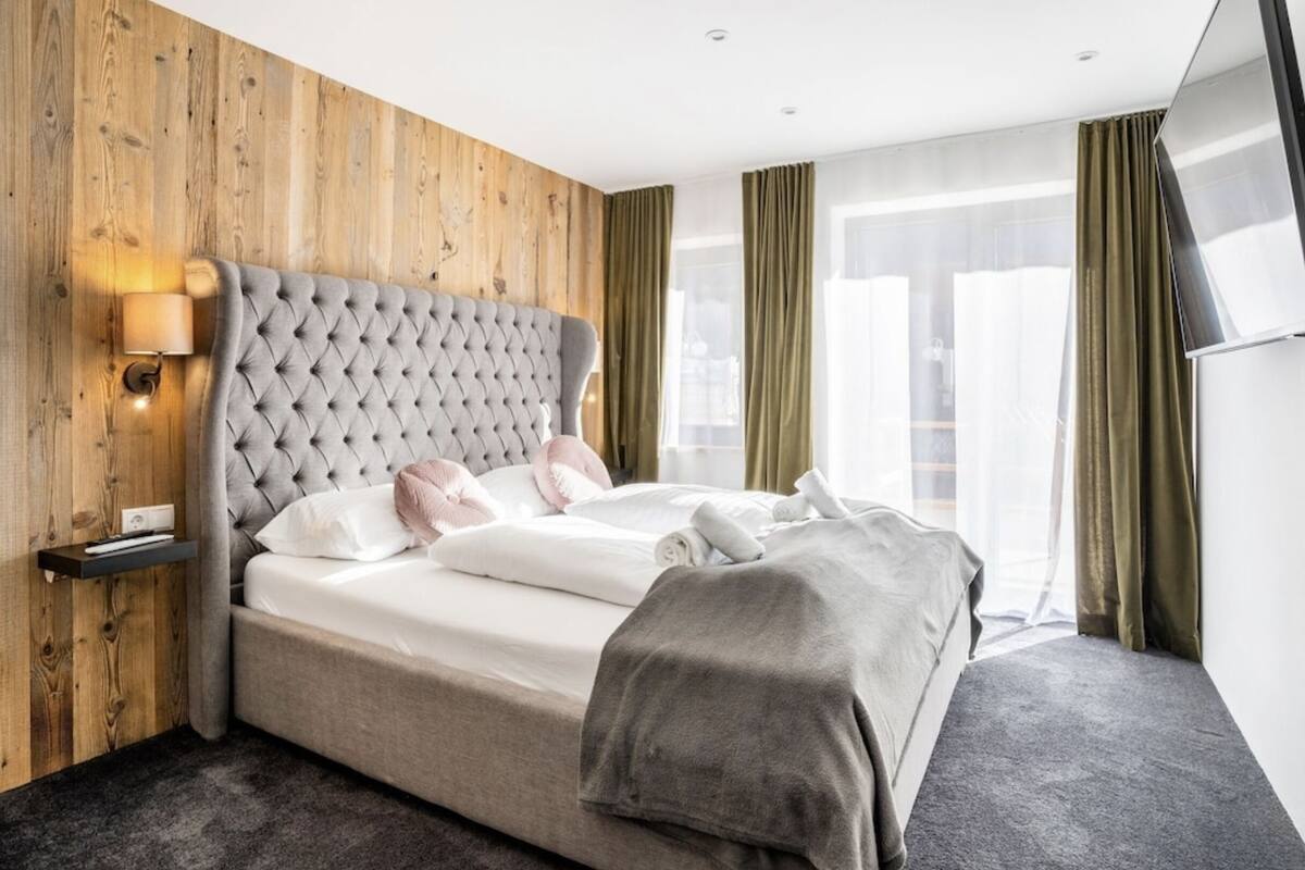 A modern hotel room with a wooden wall, a tufted headboard, a bedside table with a lamp, a large bed with pillows and a blanket, and a TV on the wall.
