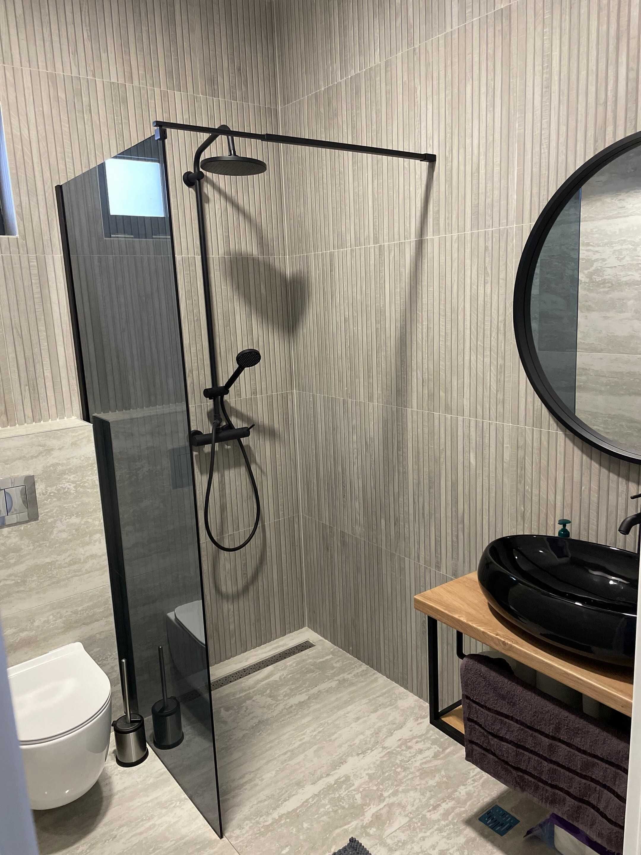 A modern bathroom with a glass-enclosed shower, a round mirror, and a wooden vanity with a black sink.