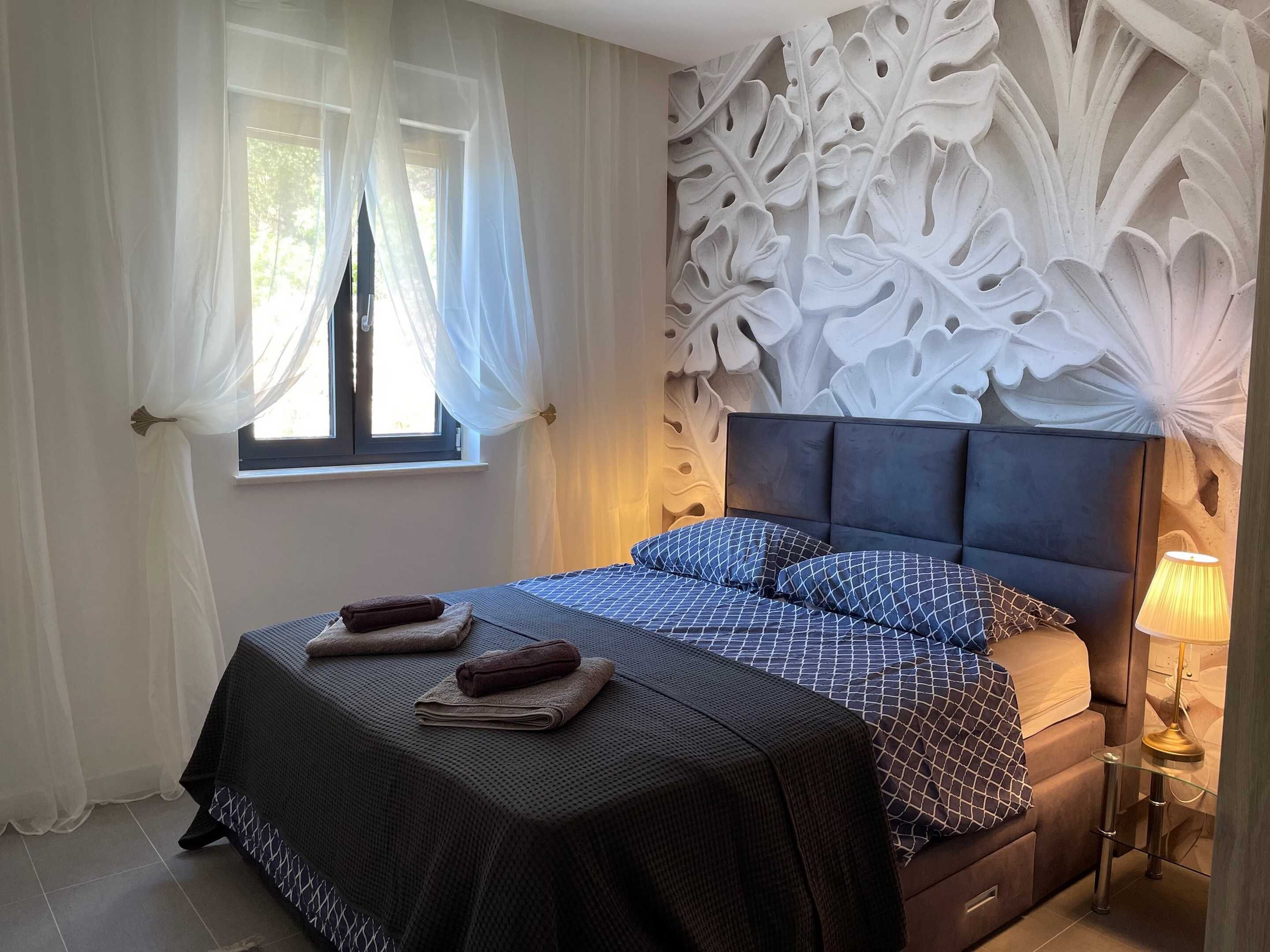 A bedroom with a bed, a window with sheer curtains, a decorative wall panel, and a bedside lamp.