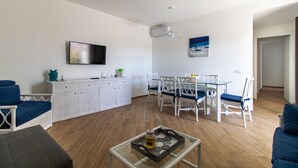 Una sala de estar moderna con una mesa de centro de vidrio, muebles blancos, un televisor de pantalla plana y un comedor con sillas blancas y una mesa de vidrio.