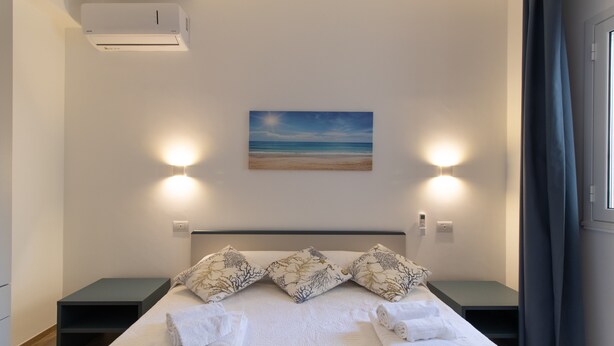 Una cama bien hecha con almohadas, una mesita de noche, un aire acondicionado de pared y un cuadro con una playa.