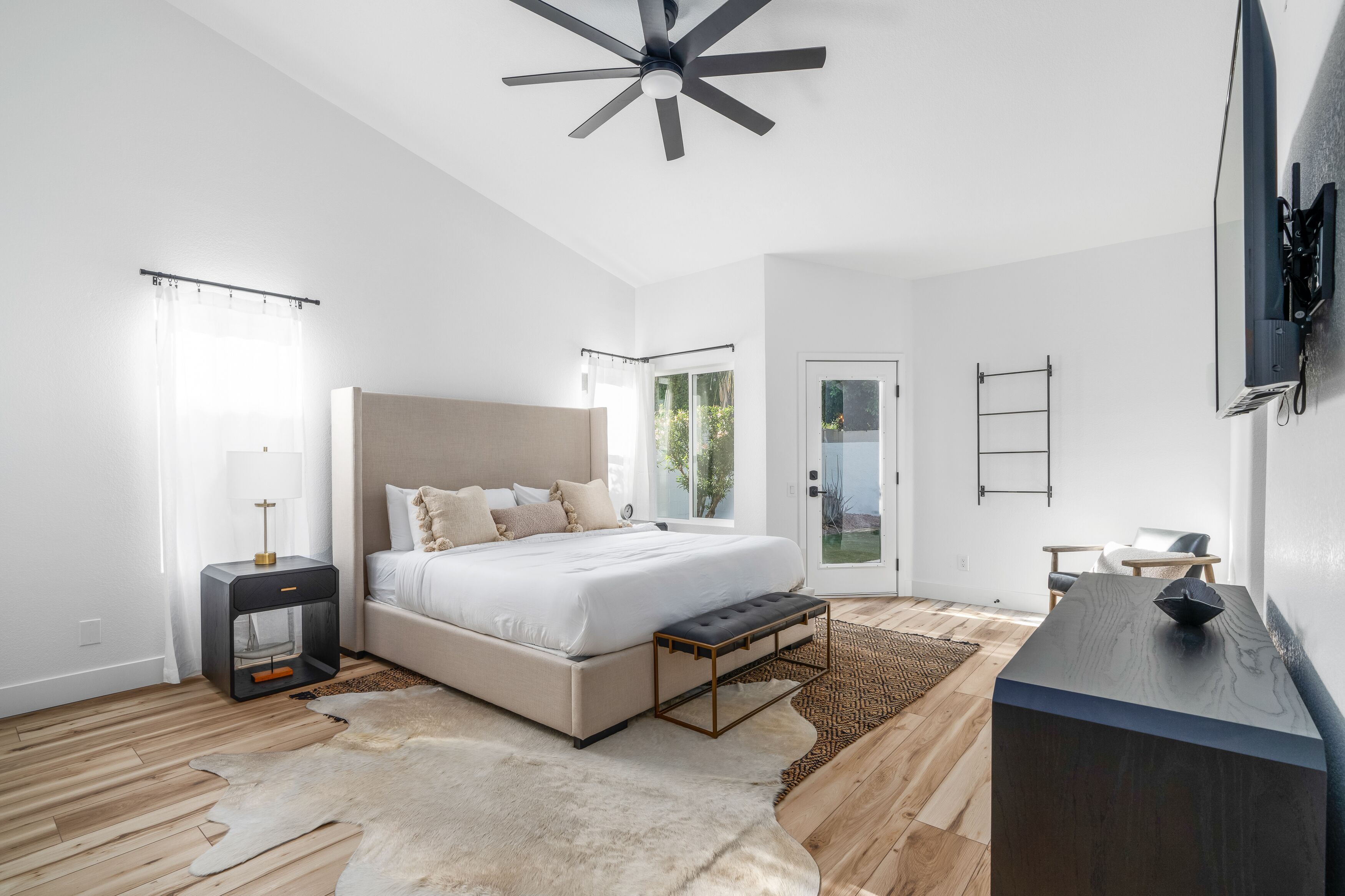 A modern bedroom with a large bed, a ceiling fan, a nightstand, a bench, and a TV mounted on the wall.