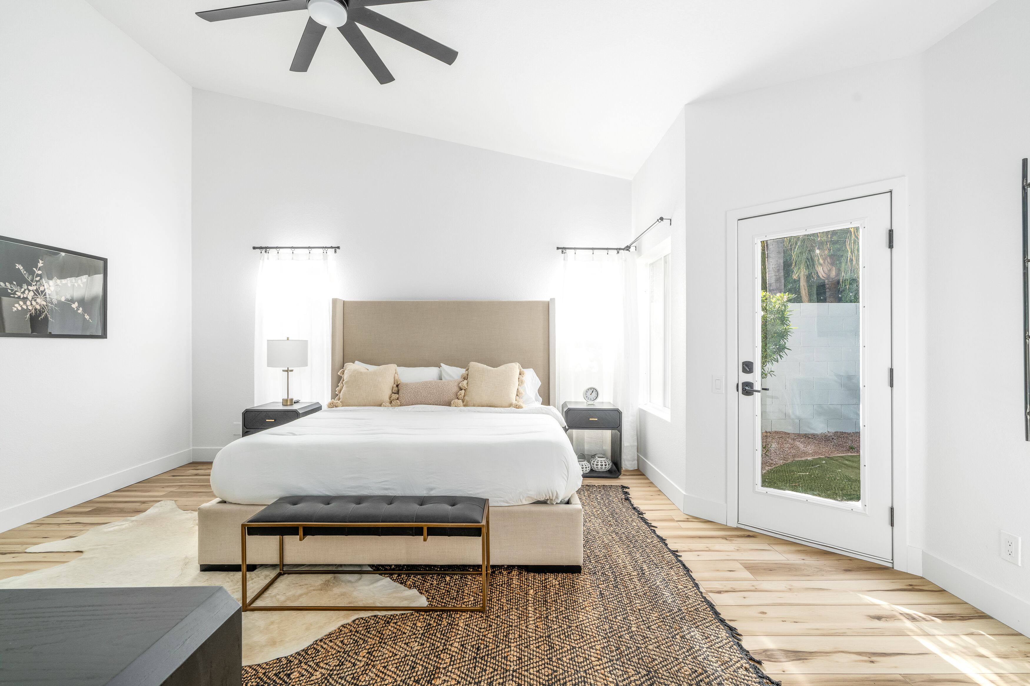 A bedroom with a large bed, bedside tables, a bench, a ceiling fan, and a door leading to a garden.