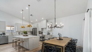 A modern kitchen with white cabinetry, a central island, pendant lighting, and a dining area with a wooden table and chairs. - Black Palm! Sparkling Pool, Pool Table, Close To Kierland Entertainment, Newly Designed Beauty! (Scottsdale)