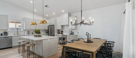 A modern kitchen with white cabinetry, a central island, pendant lighting, and a dining area with a wooden table and chairs.