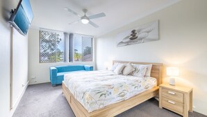 Ein Schlafzimmer mit einem Bett, einem blauen Sessel, einem Nachttisch mit Lampe, einem Deckenventilator und einem an der Wand befestigten Fernseher.