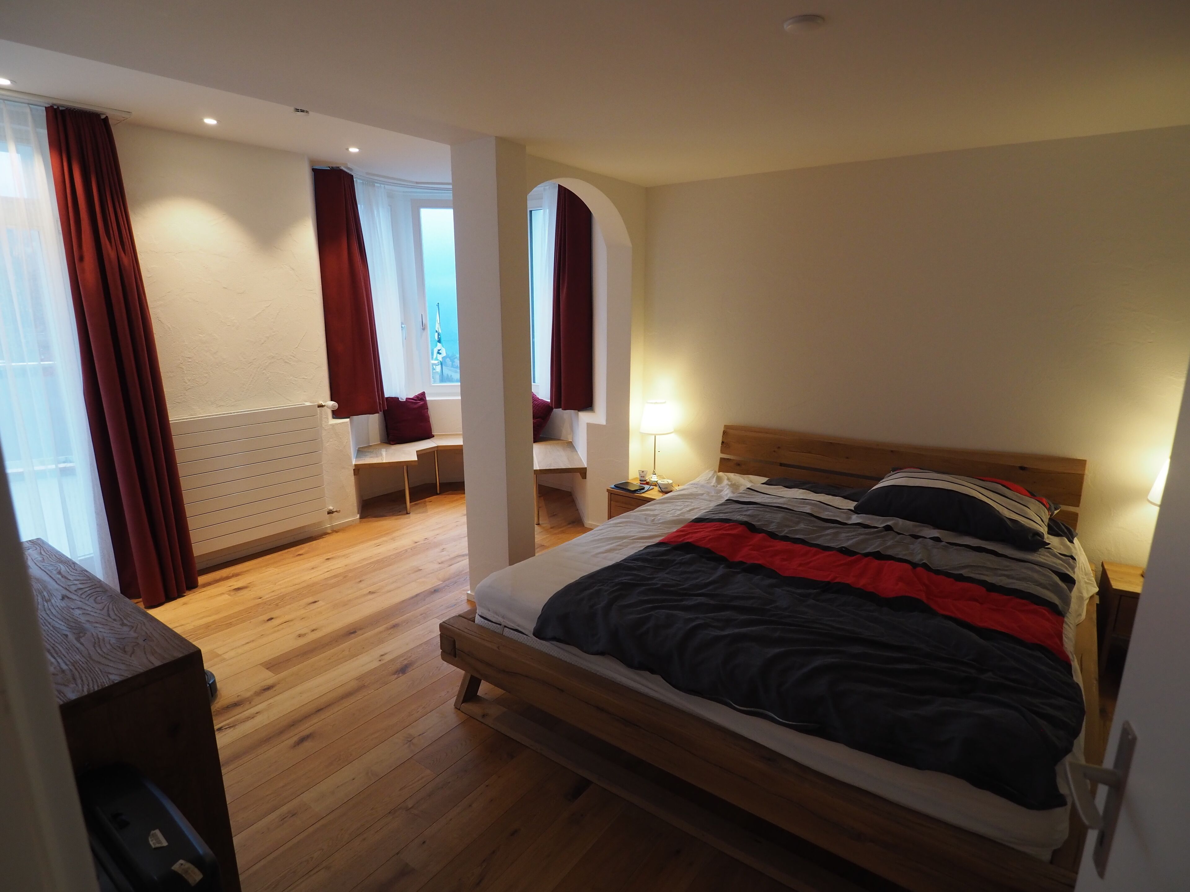 A bedroom with a wooden bed, a nightstand with a lamp, a window with curtains, and a radiator.