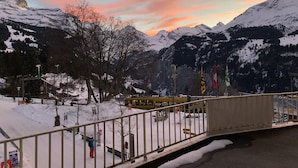 Paisaje montañoso nevado con un atardecer, un puente y algunos edificios al frente.