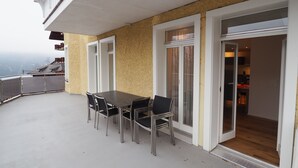 Un balcón con mesa y sillas, una puerta corrediza de vidrio que da a una cocina, y vista a un paisaje montañoso.