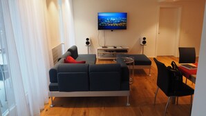 Una sala de estar moderna con un sofá, una mesa de centro y un televisor montado en la pared. Se observan dos altavoces y una pequeña mesa con un ordenador portátil.