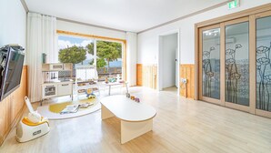 A bright room with a kitchen, a dining area, and a large window with a view of trees and a building. - Yeosu Hi Mom Kids Family Pension Gold (Yeosu)