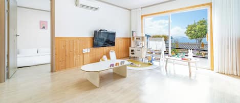A spacious living room with a wooden floor, a flat-screen TV mounted on the wall, a dining area with a table and chairs, and a large sliding glass door leading to an outdoor balcony with a view of the sea.