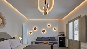 A modern hotel room with a bed, a sofa, a small table, a desk, and a window. - Mountain and Sea Suites (Santorini)