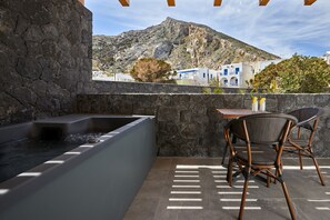 A stone-walled outdoor area with a hot tub, wicker chairs, and a view of a mountainous landscape. - Mountain and Sea Suites (Santorini)