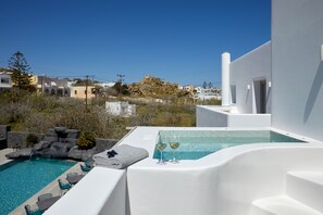 A modern outdoor pool area with a hot tub, two glasses of white wine, and a towel on a table. - Mountain and Sea Suites (Santorini)
