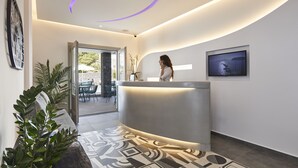 A modern reception area with a curved reception desk, a large framed artwork on the wall, and a view into the outdoor seating area. - Mountain and Sea Suites (Santorini)