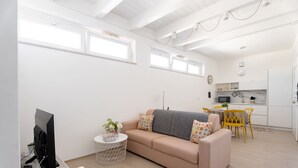 Un salon moderne avec un canapé, un téléviseur et une petite table ronde. On aperçoit, au fond, un coin cuisine avec une table à manger et des chaises.