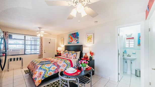A well-lit bedroom with a bed, bedside tables, a desk with chairs, a ceiling fan, and a bathroom visible through an open door. - 🏳️‍🌈 #2:  Walk to Wilton Drive (with kitchen) (fort lauderdale)