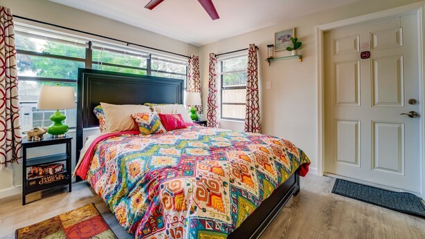 A bedroom with a colorful bedspread, a ceiling fan, a window with curtains, a bedside table with a lamp, and a door with a doormat. - 🏳️‍🌈 #3 Walk to Wilton Drive (fort lauderdale)