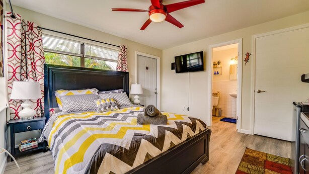 A bedroom with a bed, bedside tables, a ceiling fan, a TV, and a bathroom visible through an open door.
