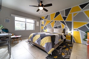 A bedroom with a bed, a desk, a chair, a ceiling fan, and a window.