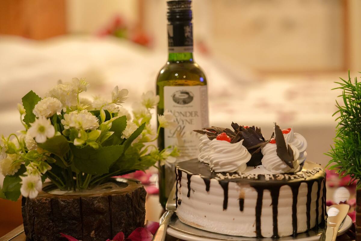 A cake with white frosting and chocolate drizzle, a bottle of wine, and a vase with white flowers on a table.