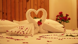 A bed with white towels arranged in a heart shape, a single rose, and scattered rose petals. - Hotel Ireland (Manali)