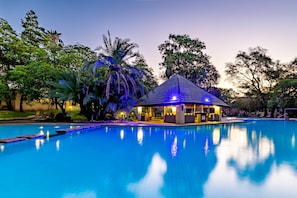 Piscina con zona de estar techada y sillones de descanso, rodeada de árboles y un edificio bien iluminado.