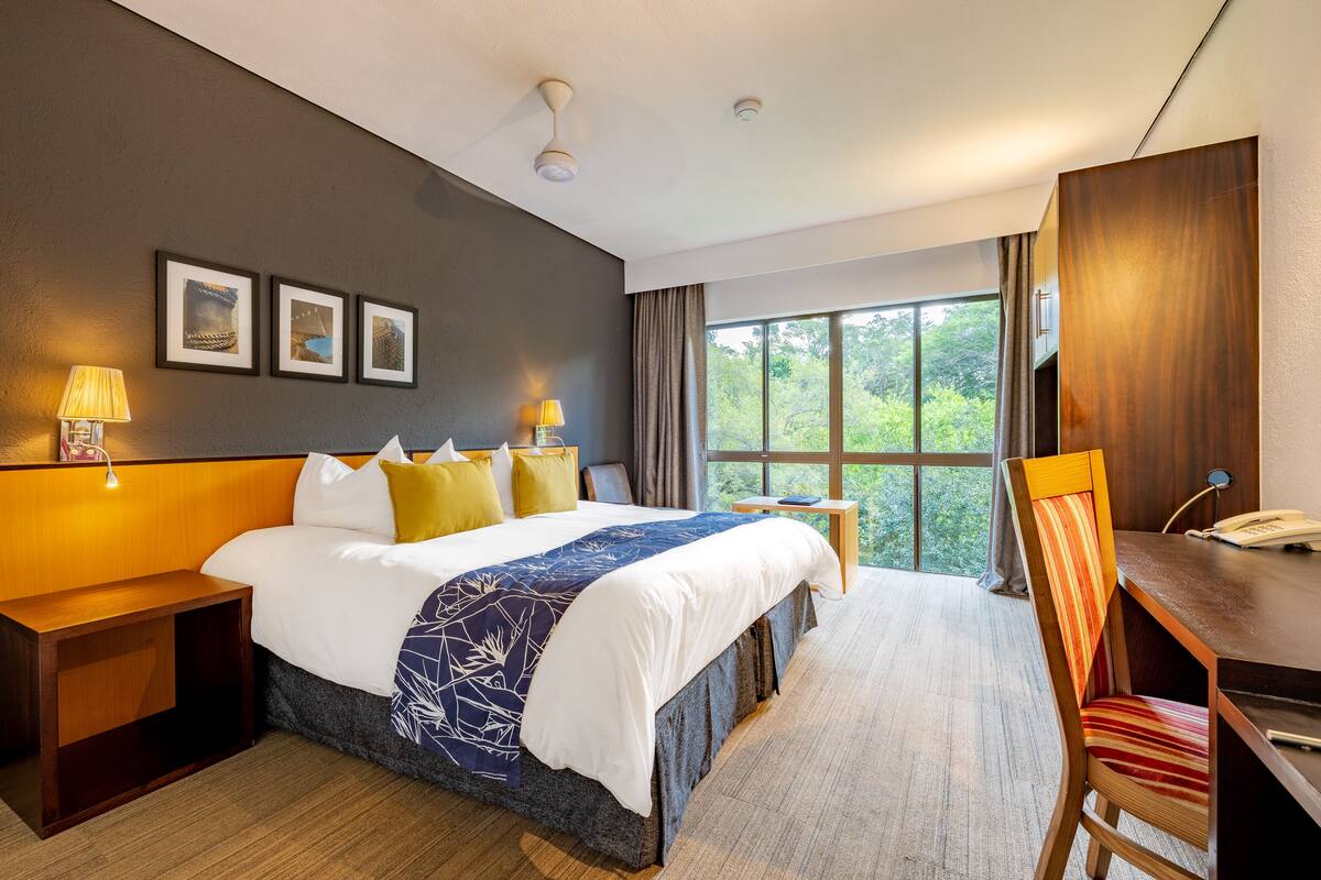 Habitación de hotel con una cama grande, lámparas de noche, un escritorio con una silla y vistas a zonas verdes por la ventana.