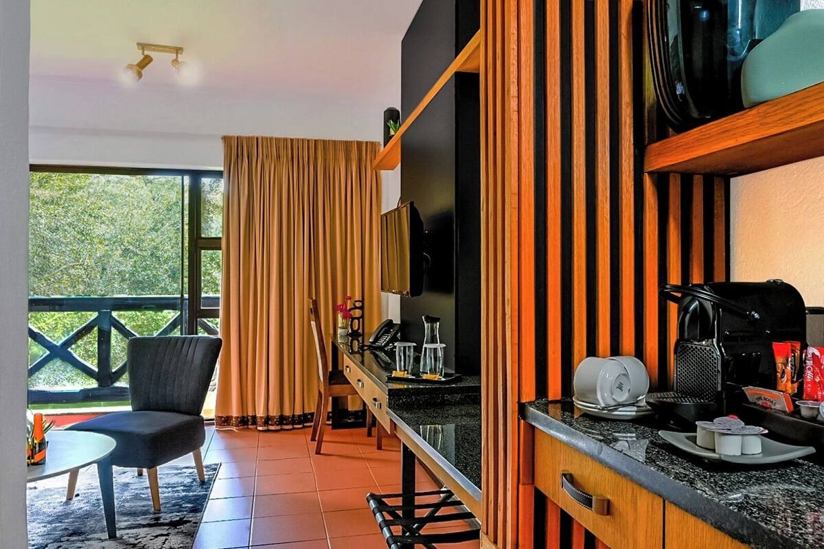 Una habitación de hotel moderna con un ventanal grande, un comedor con mesa y sillas, una kitchenette y una zona de estar con una silla y una mesita.