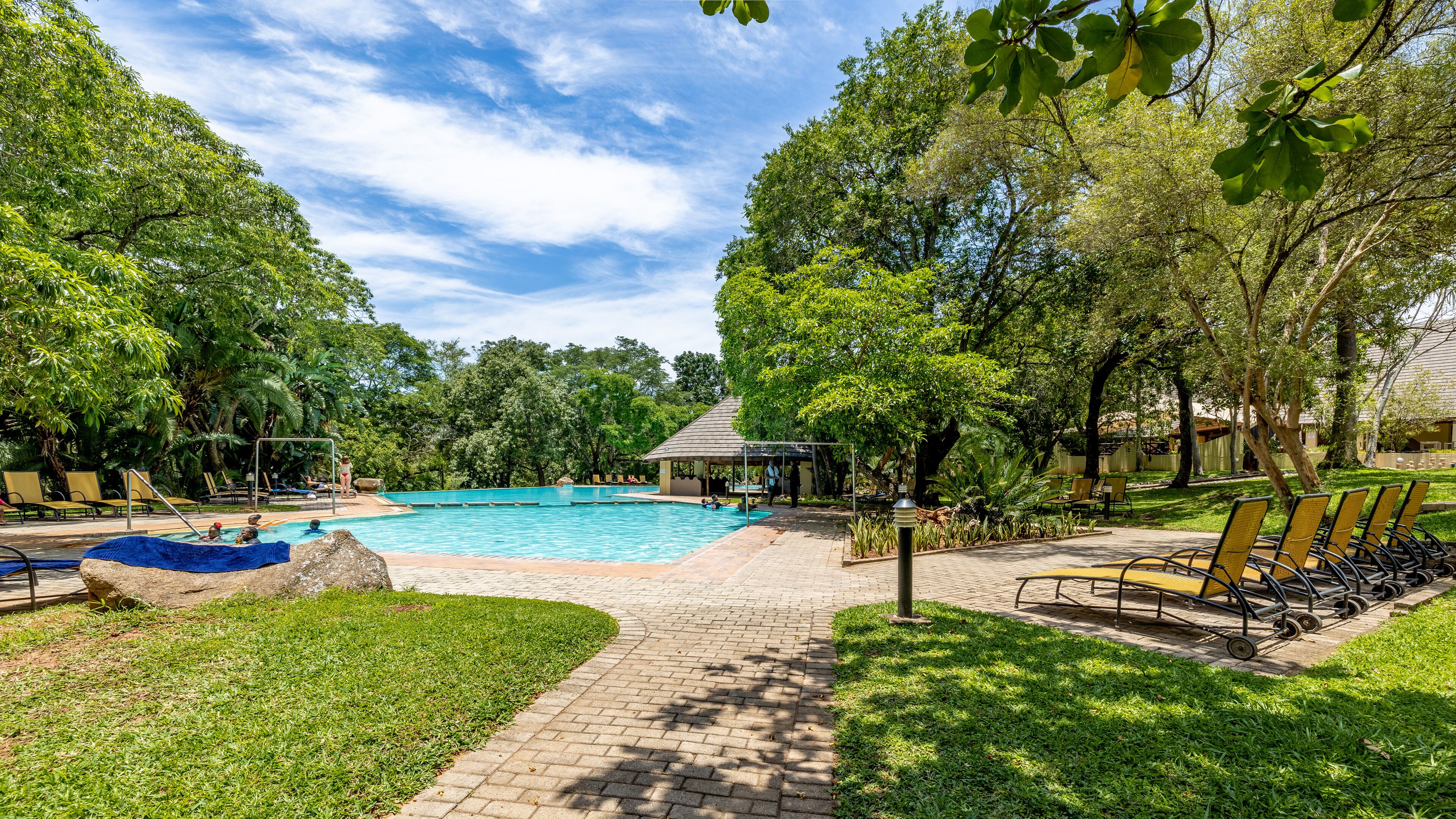 A pool area with a clear blue pool, surrounded by lush greenery and shaded by trees.