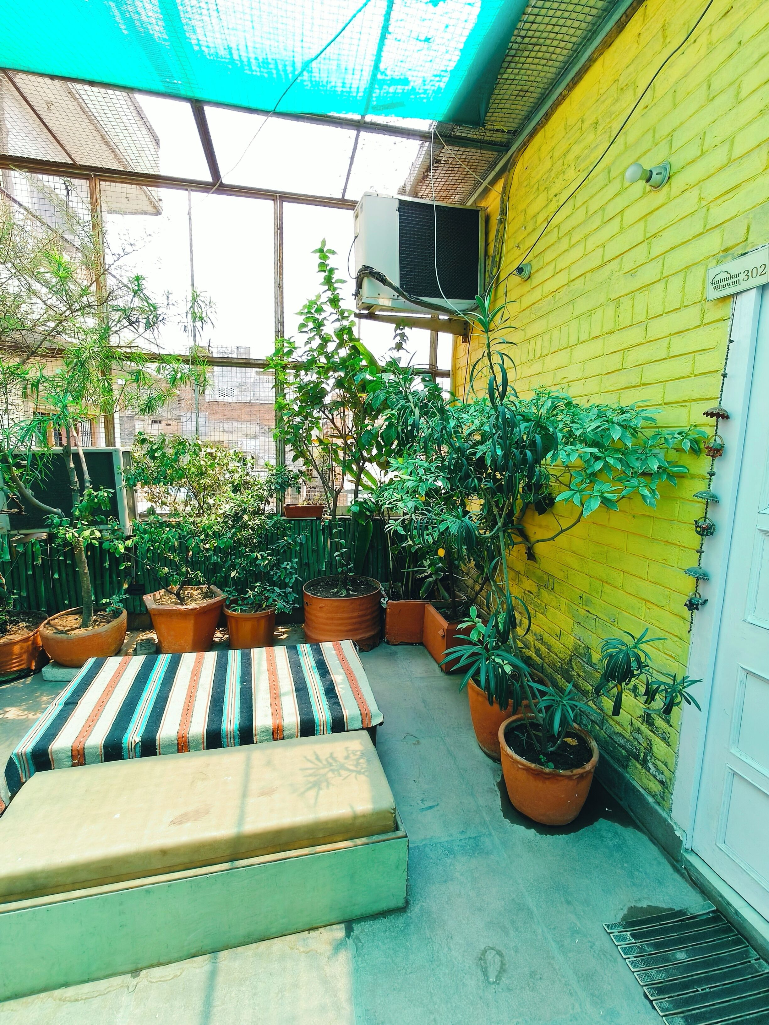 Un pequeño espacio al aire libre amurallado con un muro de ladrillo amarillo, varias plantas en macetas, un cojín a rayas y un banco.