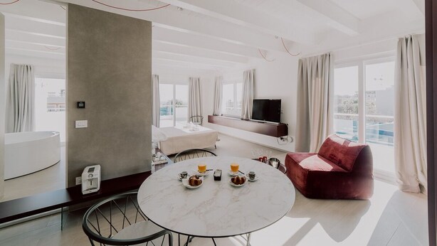 A modern hotel room with a round marble table set for breakfast, a red sofa, a TV, and a large window with curtains. - Suite Donna Edwige by MONHOLIDAY (Monopoli)