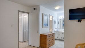 A hallway leading to a bathroom with a large mirror, a wooden chest, and a TV mounted on the wall.