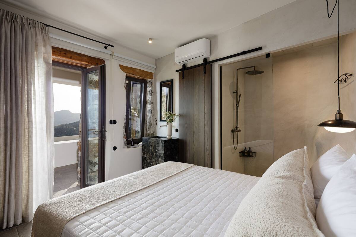A bedroom with a large bed, a sliding glass door leading to a balcony, a shower, and a mounted air conditioner.