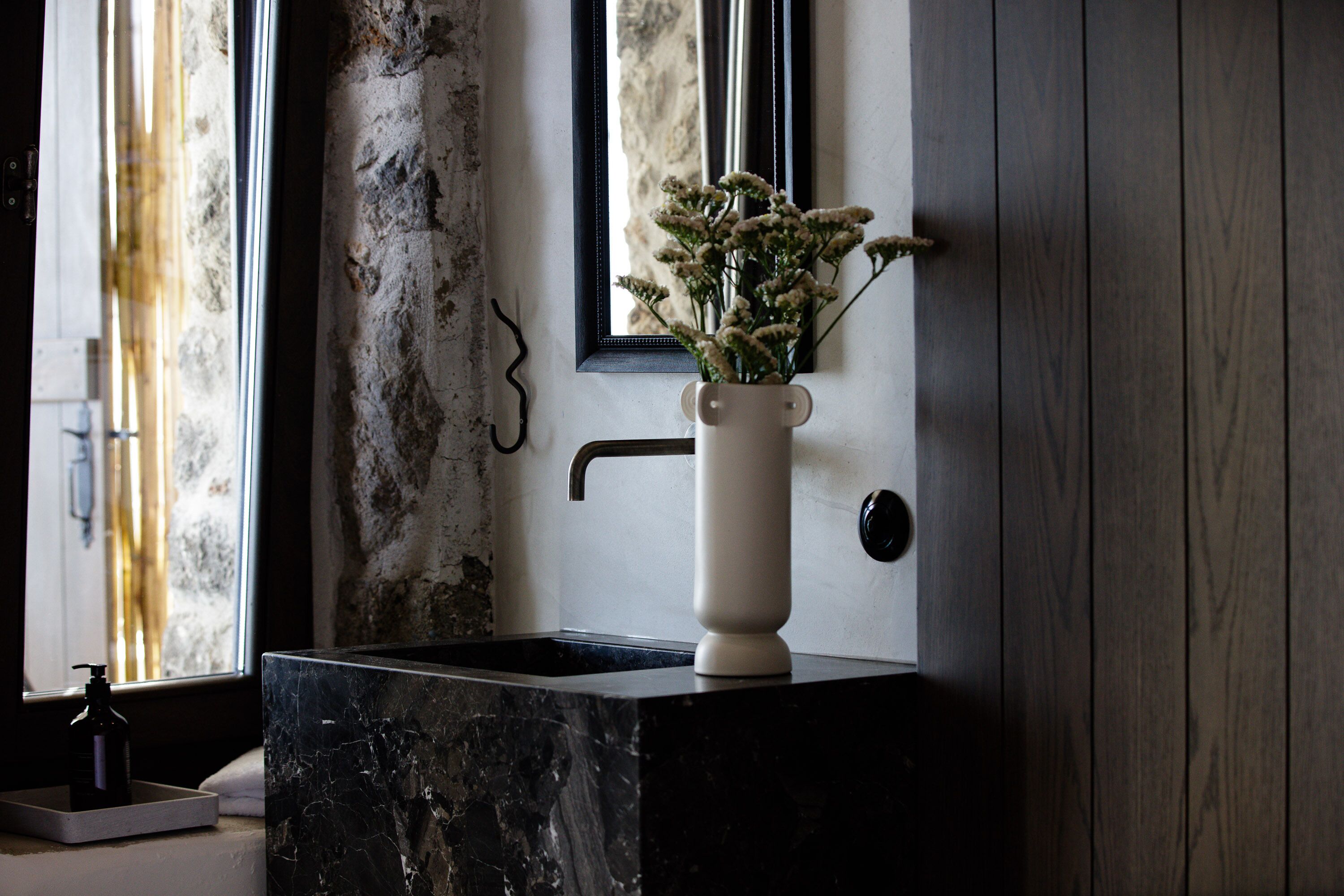 Ein Badezimmer mit Marmorwaschbecken, einer weißen Vase mit Blumen, einem Spiegel und einem Fenster mit Blick auf eine Steinwand.