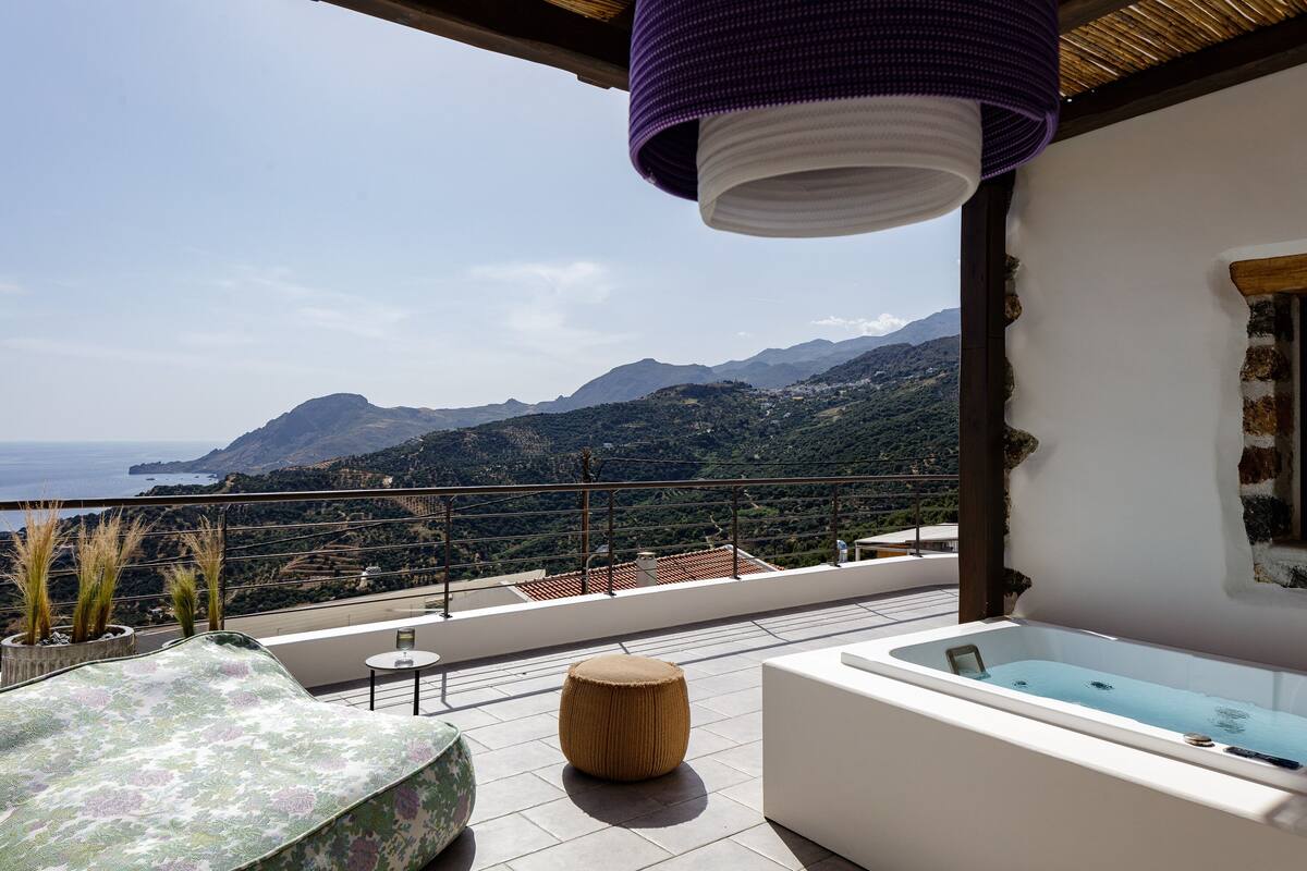 A balcony with a view of mountains and the sea, a white bathtub, and a woven ottoman.
