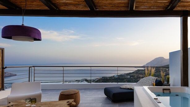 A balcony with a view of the sea, a white chair, a wooden table, and a black ottoman. - Fedra Suites (Agios Vasileios)