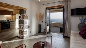 A modern living room with a stone pillar, a dining table with a cup, a sofa, and a TV. - Fedra Suites (Agios Vasileios)
