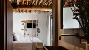 A bedroom with a bed, a wooden ceiling, a stone wall, a window, and a sink. - Fedra Suites (Agios Vasileios)