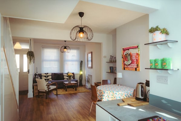 A cozy living room with a sofa, a coffee table, and a dining area with a table and chairs. There is a kitchen area with a kettle and a shelf with plants.