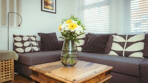 Un soggiorno con tavolino in legno, un vaso di vetro con fiori, un divano grigio con cuscini decorati e una cornice appesa al muro.